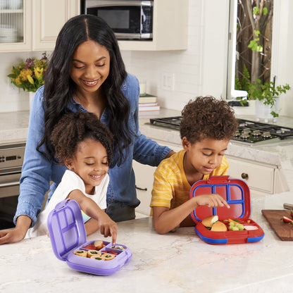 A mother smiles as she watches her young daughter and son excitedly explore their lunch options from open colorful bentgo boxes on a kitchen island.