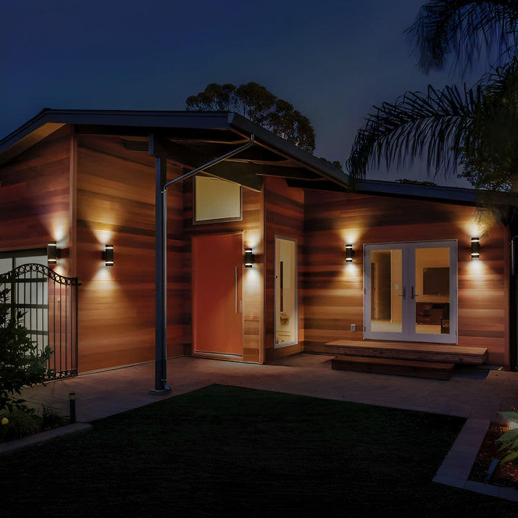 The image shows a modern home exterior at twilight, warmly lit by wall-mounted lights that cast a soft glow on the wood siding.